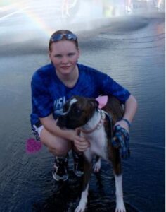 Person kneeling beside a brown and white dog on wet pavement, with a faint rainbow above.