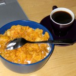 A bowl of cornflakes with milk and a spoon next to a cup of black coffee on a table.