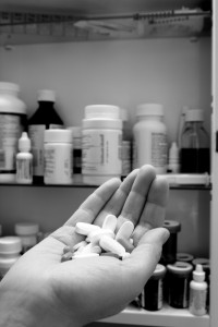Hand holding pills in front of an open medicine cabinet filled with bottles and containers.
