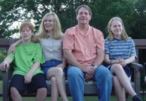 Four people sit together on an outdoor bench, smiling, with trees in the background.