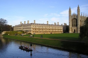 People punting on a river by historic university buildings and a large chapel on a grassy lawn.