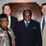 Four adults in business attire pose together and smile in front of a wooden plaque.