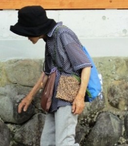 Elderly person with hat and bags walking beside a stone wall.