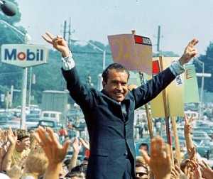 Man in a suit raises both arms in a V-sign gesture at an outdoor event, crowd and signs visible.