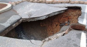 Large sinkhole in a paved road with broken asphalt and exposed earth underneath.