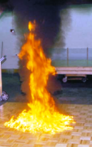 A fire burns intensely on a wooden floor, producing thick black smoke indoors.