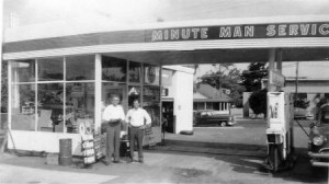Two men stand outside a vintage Minute Man Service gas station with pumps and cars visible.