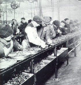 Factory workers assemble parts on an industrial production line in a historical black-and-white photo.