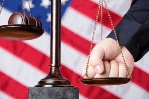A hand tips the scales of justice in front of an American flag background.