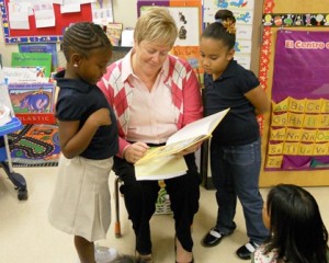 A teacher reads a book to three young students in a colorful classroom setting.