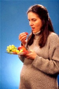 Pregnant woman eating fruit from a plate against a blue background.