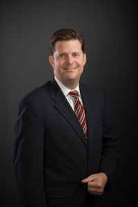 Man in dark suit and striped tie poses against a plain dark background, facing the camera.