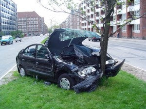 A black car crashed head-on into a tree on a grassy area beside a city street.