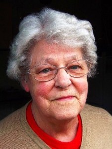 Elderly woman with gray hair and glasses, wearing a red top and beige sweater, facing camera.