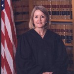A judge in black robes sits in front of bookshelves and an American flag.