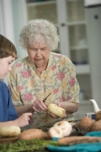 Elderly woman peeling a potato with a child beside her in a kitchen setting.