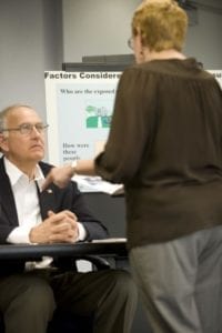 A seated man listens as a standing woman gestures and speaks in a meeting room.