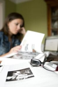 A woman reads a book at a desk with papers, a laptop, and eyeglasses in the foreground.