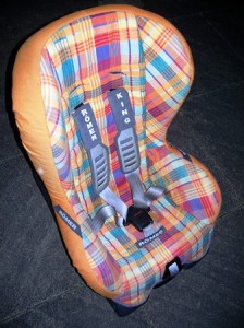 A plaid-patterned child car seat with black harness straps on a dark floor.