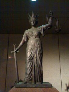 Bronze statue of Lady Justice holding scales and a sword, standing on a pedestal indoors.