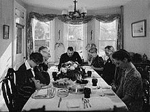 Eight people sit around a dining table with heads bowed, appearing to pray before a meal.