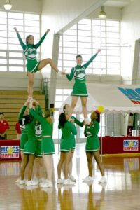 Cheerleaders in green uniforms perform a stunt formation inside a gymnasium.