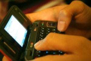 Hands typing on a small, black flip phone with a physical keyboard and lit screen.