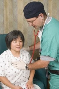 Doctor uses stethoscope to examine a seated patient wearing a hospital gown.