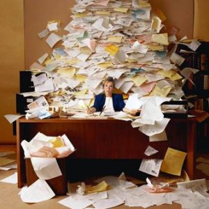 Person sitting at a desk overwhelmed by a large pile of scattered papers and documents.