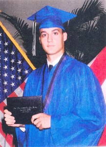 Young man in blue graduation gown and cap holding a diploma, standing by flags.