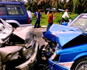 Two cars with severe front-end damage after a head-on collision on a road, people nearby.
