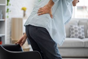 Person holding lower back in pain while standing up from a chair in a living room.