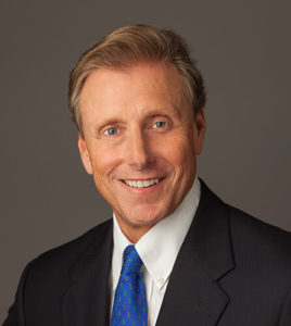 Smiling man in a suit and blue tie against a plain gray background.