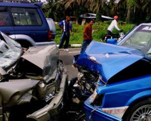 Two heavily damaged cars after a head-on collision with bystanders and a motorcyclist nearby.