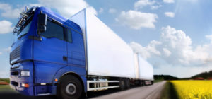 Blue semi-truck with a white trailer driving on a rural road beside a yellow field and blue sky.