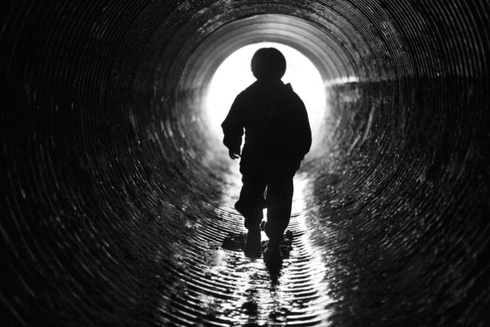 contrasty black and white image of silhouette of a child walking down the inside of a large drainage pipe.