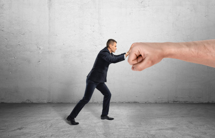 Businessman trying to resist a huge male fist and move it away on a grey background. Containing market pressure. Being competitive. Protecting a small business.