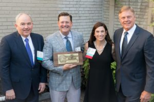 Four people in formal attire pose with a plaque in front of a brick wall outdoors.
