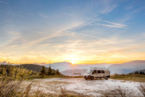A camper van is parked near hazardous refrigerators on a hilltop at sunrise, mountains behind.