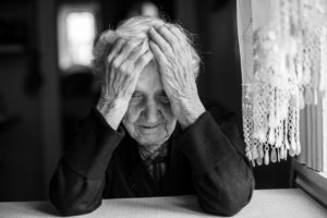 Elderly woman in a nursing home sits at a table, holding her head in her hands, looking down.