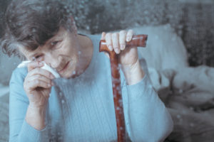 Elderly woman wipes her eye with a tissue while holding a cane, seen through a rainy window.