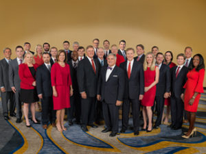 A group of professionally dressed men and women pose together against a gold background.