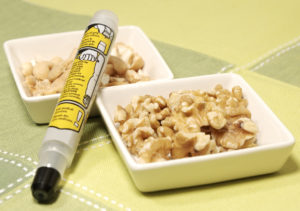 A food allergy injector next to small bowls of walnuts and other nuts on a table.