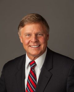 Smiling man in a dark suit, white shirt, and striped red tie against a plain background.
