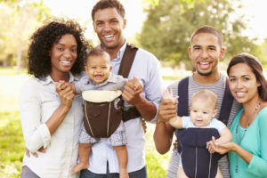 Two couples with babies in baby slings, smiling together outdoors in a sunny park.