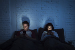 Man and woman in bed at night, both lost in texting addiction on their smartphones.