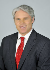 Man in a dark suit and red tie smiling at the camera against a plain background.