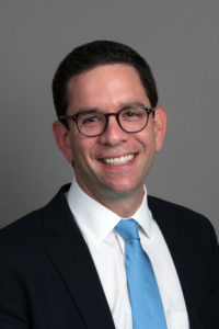 Man in suit, white shirt, and blue tie smiling in front of a plain gray background.