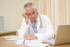 Male doctor in white coat sits at desk looking at laptop with paperwork in front of him.