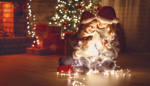 Child and adult in Santa hats sit by a Christmas tree, playing with string lights.